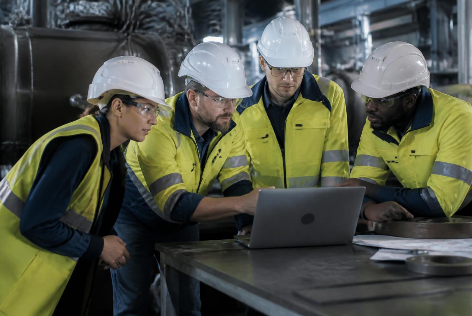 Equipe multidisciplinar reunida em torno de um notebook dentro de uma planta industrial